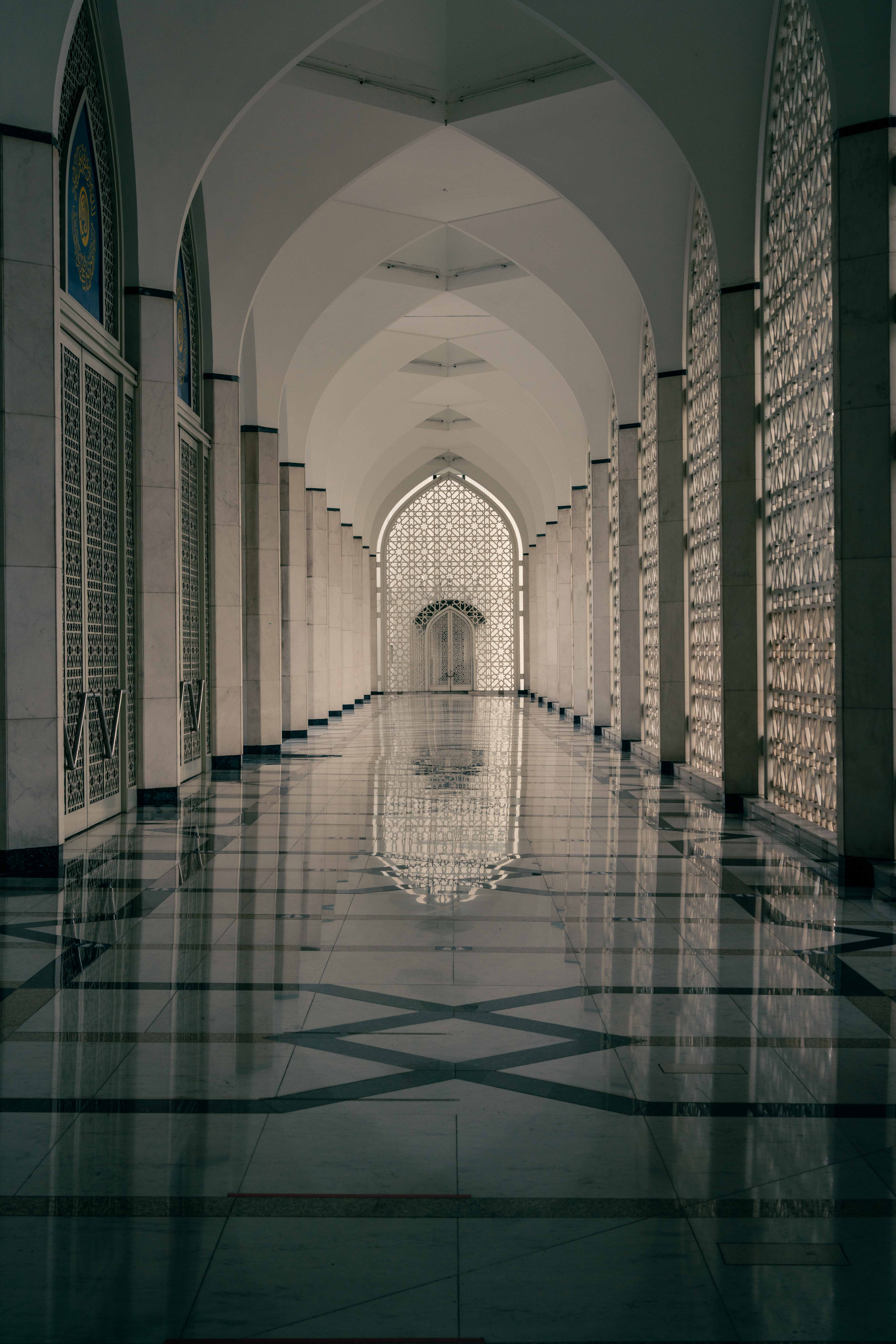A vertical interior corridor with repeating white pointed arches, patterned screens on both sides, a bright lattice doorway at the far end, and glossy floor reflections.